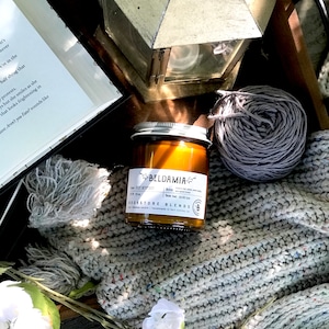Peut inclure: Nature morte avec une bougie allumée dans un bocal en verre, une couverture en tricot grise, une pelote de laine grise, une lanterne vintage et un livre. L'étiquette de la bougie indique "BELDAMIA BOOKSTORE BLENDS".