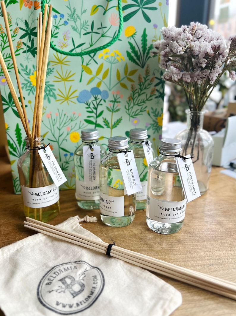 May include: Five clear glass bottles with labels and wooden sticks in a white bag. The bottles are filled with a clear liquid and have labels that say "The Orangery", "The Grangery", "Magic Shop", "Garden Moon", and "Beldamia Reed Diffuser".
