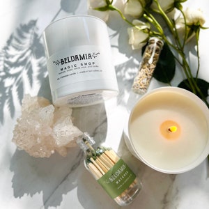 May include: A white candle with a black label that reads "BELDAMIA MAGIC SHOP incense, violet, white rose, and tonka soy + beeswax candle | made in fort collins, co". The candle is sitting on a white surface with a small bottle of matches and a bouquet of white roses.