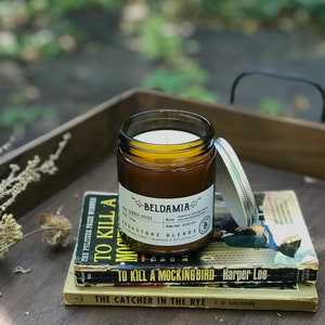May include: A brown glass candle jar with a white label that reads "BELDAMIA" and "BOOKSTORE BLENDS". The candle is sitting on a stack of three books: "To Kill a Mockingbird", "The Catcher in the Rye", and a book with the title partially obscured.
