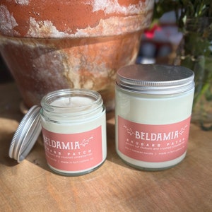May include: Two glass jars with white lids and pink labels. The labels say "BELDAMIA" and "RHUBARB PATCH". The jars are filled with white wax and are likely candles.