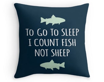 May include: A square dark blue throw pillow featuring two light green fish silhouettes and the white text "TO GO TO SLEEP I COUNT FISH NOT SHEEP" centered between the fish graphics. The design is printed on a solid dark blue fabric cover.