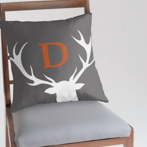 May include: A square throw pillow in dark gray featuring a white silhouette of a deer head with large antlers. Centered between the antlers is a large, bold, orange capital letter D. The pillow is positioned on a wooden chair with a light gray seat.