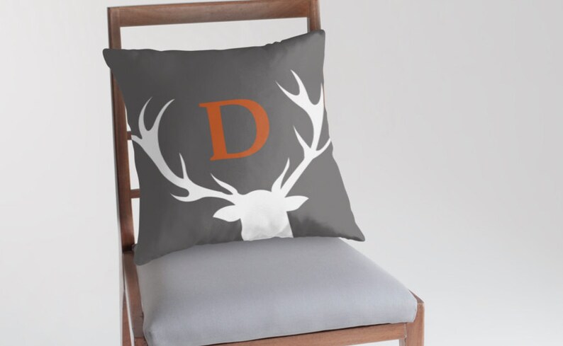 May include: A square throw pillow in dark gray featuring a white silhouette of a deer head with large antlers. Centered between the antlers is a large, bold, orange capital letter D. The pillow is positioned on a wooden chair with a light gray seat.