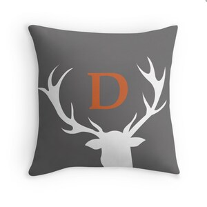 May include: A square throw pillow in dark gray featuring a white silhouette of a deer head with large antlers. Centered between the antlers is a large, bold, orange capital letter D. The design is printed on the front of the fabric cover.