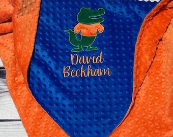 May include: A soft, textured minky blanket featuring a bright blue center panel and vibrant orange borders. The center displays an embroidered green alligator wearing an orange shirt, with the name David Beckham stitched in orange cursive text below.