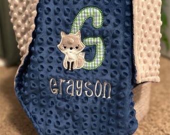 May include: A navy blue baby blanket with a plush, dimpled texture. The blanket features an embroidered fox and the name "Grayson" in silver thread. A large green and white plaid "G" is also embroidered on the blanket. The blanket has a soft, tan, bumpy border.