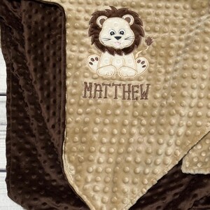 May include: A soft, two-toned baby blanket with a brown and tan color scheme. The blanket features a cute lion applique with the name "MATTHEW" embroidered below. The blanket has a textured, dotted surface.