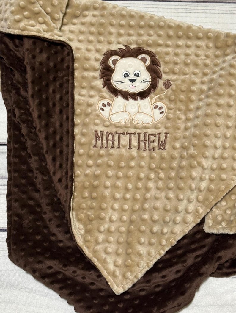 May include: A soft, two-toned baby blanket with a brown and tan color scheme. The blanket features a cute lion applique with the name "MATTHEW" embroidered below. The blanket has a textured, dotted surface.
