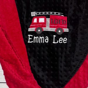 May include: A black and red baby blanket with a fire truck design. The fire truck is red with silver accents and the letters "LGFD". The name "Emma Lee" is embroidered in white.