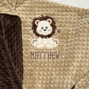 May include: A tan and brown baby blanket with a lion applique. The lion is beige with a brown mane and tail. The name "MATTHEW" is embroidered below the lion in brown thread. The blanket has a textured, dotted pattern.