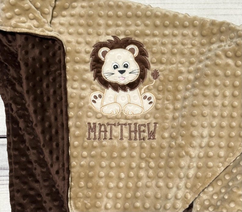 May include: A tan and brown baby blanket with a lion applique. The lion is beige with a brown mane and tail. The name "MATTHEW" is embroidered below the lion in brown thread. The blanket has a textured, dotted pattern.