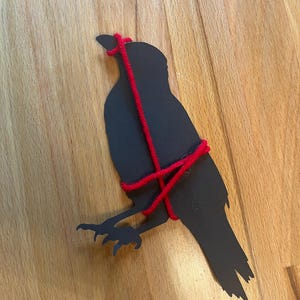 May include: A flat, black silhouette of a bird, specifically a crow or raven, wrapped in thick, bright red yarn. The yarn is wound around the bird's neck and body in a crisscross pattern, creating a stark contrast against the dark cutout shape.