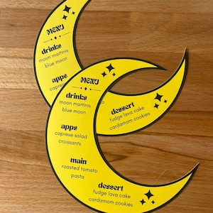 May include: Two crescent moon-shaped yellow paper menus with black text and star icons. The menus list sections for drinks, apps, main, and dessert, including items like moon martinis, blue moon, caprese salad, croissants, roasted tomato pasta, fudge lava cake, and cardamom cookies.