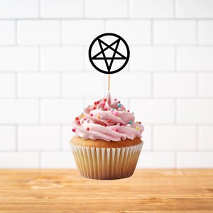May include: A cupcake with light pink frosting and multicolored sprinkles topped with a black circular pentagram topper on a thin wooden pick. The cupcake sits in a white paper liner, showcasing a golden-brown cake base and swirl of icing.