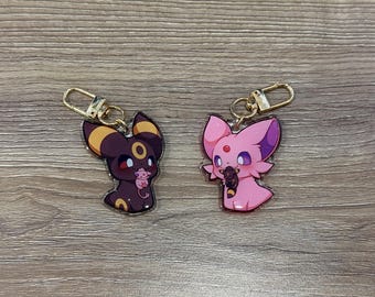 May include: Two cartoon character keychains. One is dark brown with yellow accents, the other is pink with purple accents. Both have gold-colored key rings and clasps. The characters are holding smaller versions of themselves.