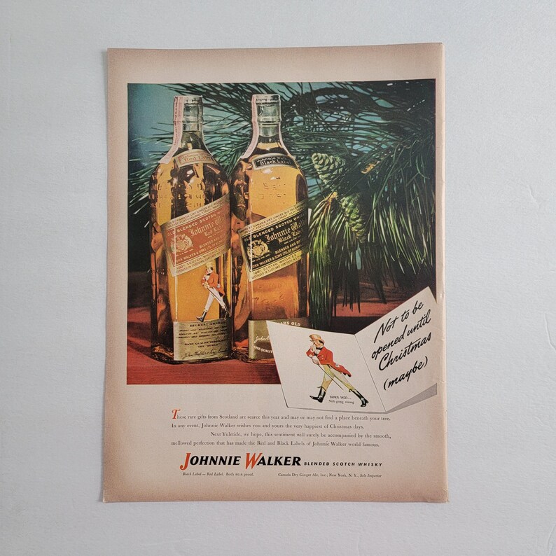 May include: Vintage advertisement featuring two bottles of Johnnie Walker Blended Scotch Whisky. The bottles have red and black labels. A small illustration of a man in a red coat is on the labels. Text reads "Not to be opened until Christmas (maybe)".