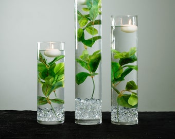May include: Three clear glass cylinder vases with floating candles and green leafy stems. The vases are filled with water and clear glass beads.
