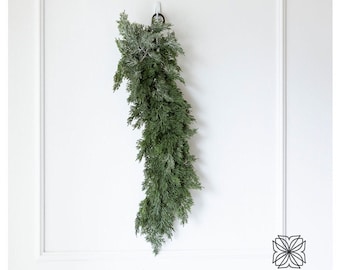 Könnte beinhalten: Ein grüner Kranz aus Zedernzweigen hängt an einem weißen Haken an einer weißen Wand mit weißen Leisten.