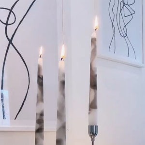 Pode incluir: Três castiçais prateados com velas marmorizadas brancas e cinzas. As velas estão acesas e os castiçais têm um design simples e moderno.