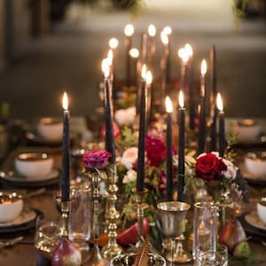 Puede incluir: Una mesa rústica con velas negras de taper, candelabros dorados y un tazón blanco con una pluma sobre un plato con estampado blanco y negro. La mesa está decorada con flores rojas y blancas y fruta.
