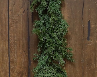 May include: A long, green garland of artificial cedar branches hangs against a woodgrain background. The garland is perfect for adding a touch of greenery to your home decor.