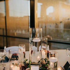 May include: A white table number card with the text "Table 4" on a white table with a centerpiece of white and pink roses, greenery, and candles in glass cylinders.