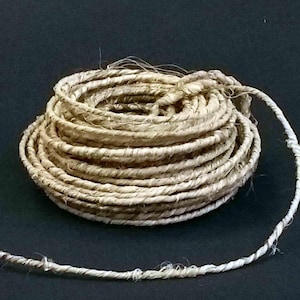 May include: A coiled rope made from natural fibres, possibly hemp or jute, with a lighter, thinner strand extending from the coil. The rope is a light tan colour and has a slightly rough texture. The background is black.