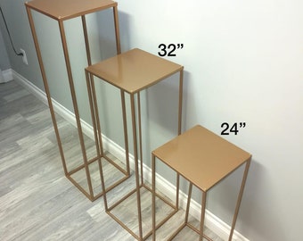 May include: Three gold metal stands of different heights, 40 inches, 32 inches, and 24 inches. The stands are square and have a simple, modern design.