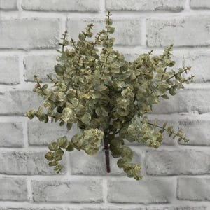 May include: A faux eucalyptus plant stem featuring multiple branches with small, rounded, sage green leaves. The foliage has a muted, dusty appearance with subtle brown undertones on the stems and leaf edges, arranged in a dense, bushy cluster.