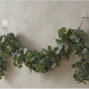 May include: A decorative garland featuring artificial eucalyptus leaves in various shades of green and muted sage. The foliage is arranged in a dense, trailing vine structure with small, light-colored buds interspersed throughout the greenery.