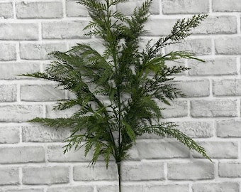 May include: A single artificial cedar branch featuring a long, thin central stem with multiple spreading, feathery green foliage sprays. The branch displays a realistic texture with fine, needle-like leaves arranged in a dense, layered pattern.