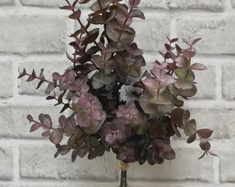May include: A bundle of faux eucalyptus leaves in shades of burgundy, purple, and green. The leaves are tied together with twine and metal stems. The background is a white brick wall.