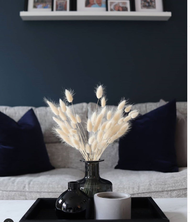 May include: A bouquet of dried pampas grass in a dark green glass vase, with two black vases and a gray concrete cylinder on a black tray. The tray is on a white surface.