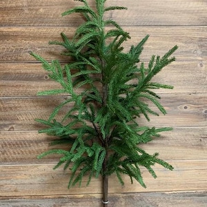May include: A green artificial pine branch with a brown stem. The branch is against a wooden background.