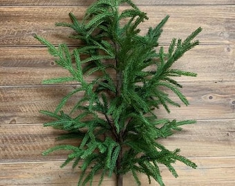 May include: A green artificial pine branch with a brown stem. The branch is against a wooden background.