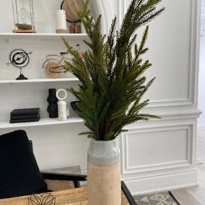 May include: A tall, light brown ceramic vase with a light gray band around the middle. The vase is filled with a green artificial pine sprig.