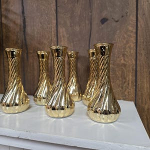 May include: A collection of seven gold-colored glass vases with a twisted, ribbed design. The vases have a bulbous base and a narrow neck, reflecting light. They are arranged on a white surface against a wooden background.