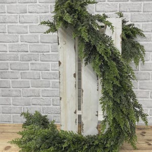 May include: A lush, artificial cedar garland featuring dense, dark green foliage with realistic needle textures. The garland is draped over a tall, weathered white wooden crate with visible distressed paint, creating a cascading effect.