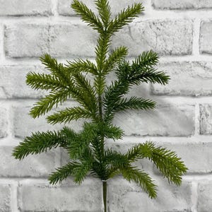 May include: A single artificial evergreen branch featuring a central stem with multiple smaller offshoots covered in short, needle-like green foliage. The branch has a realistic appearance with varying shades of green and a thin, dark brown stem base.