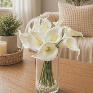 Puede incluir: Un jarrón de cristal transparente lleno de calas blancas y pequeñas piedras blancas. Las flores son de color blanco cremoso con centros amarillos y tallos verdes. El jarrón está sobre una mesa de madera.