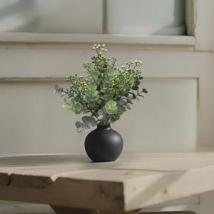 May include: A small, round, matte black vase holding a bouquet of green eucalyptus leaves and small white buds. The vase sits on a light-coloured wooden surface, with a white background.