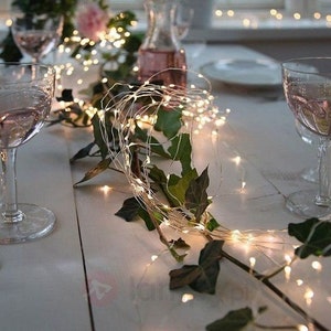 May include: A string of warm white fairy lights draped over a table setting with greenery. The lights are strung on a thin wire and create a warm, inviting ambiance.