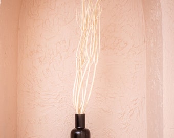 May include: A black ceramic vase with a tall, slender shape holds a bouquet of white dried branches. The vase and branches are set against a textured, light pink wall.