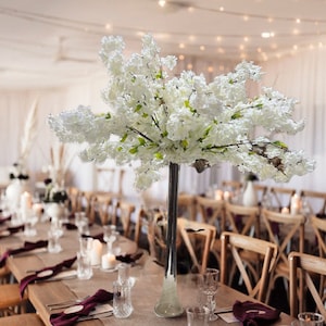 May include: A long, clear glass vase filled with white cherry blossom branches sits on a wooden table with a brown runner. The table is set with white plates, clear glasses, and burgundy napkins. The background features a white ceiling with string lights and a white wall.