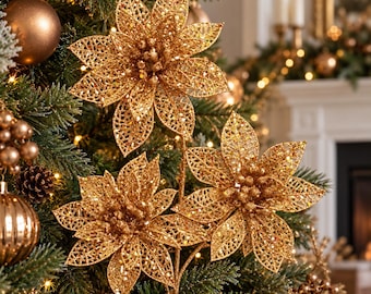 May include: A decorative floral stem featuring three gold glittered poinsettia blossoms with intricate openwork petal designs and textured centres, positioned among green pine branches and round metallic ornaments.