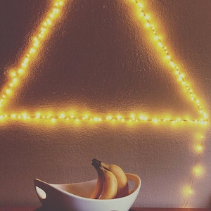 May include: A white ceramic bowl filled with yellow bananas, sitting on a table in front of a wall with a triangle of warm white string lights.