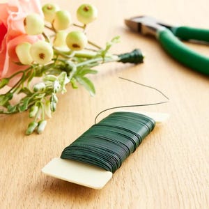 May include: A spool of dark green floral wire with a white plastic base sits on a wooden surface. A small bouquet of pink flowers and green berries is in the background, along with green handled wire cutters.