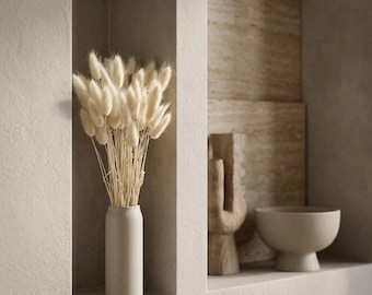 May include: A cylindrical beige vase containing a bouquet of dried cream-colored lagurus ovatus grass, also known as bunny tails. The vase is positioned in a recessed wall niche next to a wooden sculpture and a beige pedestal bowl.