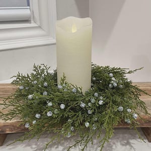 May include: A tall, off-white LED pillar candle with a wavy top edge sits in the center of a circular wreath made of green cedar-like foliage and small, frosted white berries. The candle emits a soft, warm glow from its faux flame tip.